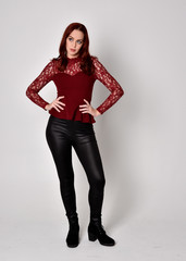 Portrait of a pretty girl with red hair wearing leather pants and long sleeved lace shirt.  full length standing pose, isolated against a studio background.