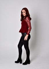 Fototapeta premium Portrait of a pretty girl with red hair wearing leather pants and long sleeved lace shirt. full length standing pose, isolated against a studio background.