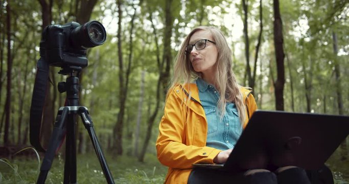 Beautiful woman with blond hair sitting near tripod with digital camera and holding portable laptop on knees. Female photographer in yellow jacket and eyeglasses working with green nature around.