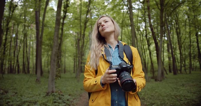 Attractive photographer with long blond hair walking among green trees with professional digital camera. Mature woman in yellow jacket taking pictures of green forest