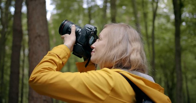 Happy photographer with blond hair taking pictures of beautiful green nature surrounding her. Mature woman in yellow jacket spending shooting time at spring forest