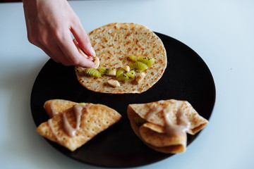 Woman's hand decorates pancakes with a fruits kiwi and banana. Black plate, white table