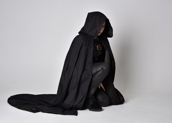 Fantasy portrait of a woman with red hair wearing dark leather assassin costume with long black cloak. full length kneeling pose, isolated against a studio background.