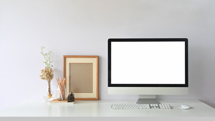 Computer monitor with white blank screen putting on workspace that surrounded by wireless mouse, keyboard, empty picture frame, pencils in glass vase and wild grass in bottle.