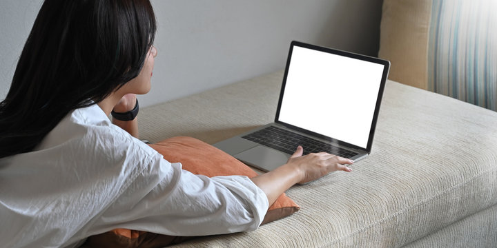Photo of young woman laying on sofa and watching/using white blank screen computer laptop over comfortable living room as background. Woman with relaxing time concept.