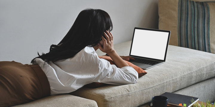 Photo of young woman laying on sofa and watching/using white blank screen computer laptop over comfortable living room as background. Woman with relaxing time concept.