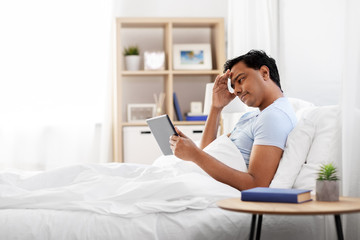 people, bedtime and rest concept - stressed indian man with tablet pc computer lying in bed at home