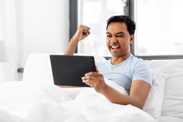 people, bedtime and rest concept - happy smiling indian man with tablet pc computer lying in bed and making winning gesture at home