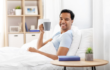 people, bedtime and rest concept - happy smiling indian man drinking coffee in bed at home
