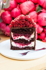 Piece of chocolate cake with cream cheese and berry filling and fresh raspberries on top. Peonies bouquet on background.