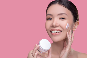 Obraz na płótnie Canvas Pretty asian girl applying cream on her face and smiling