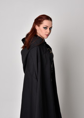 Naklejka premium Close up fantasy portrait of a woman with red hair wearing dark leather assassin costume with long black cloak. isolated against a studio background.