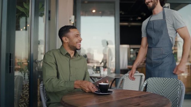 Young happy caucasian waiter serving coffee to african customer sitting in outdoor seating using digital tablet
