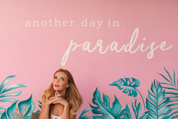 Pretty young blond girl sitting on pink wall background with tropical palm leaves and sign "another day in paradise'. Tropical summer vacation concept