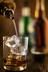 In the bar, whiskey is poured into a glass with ice, in the background there are bottles on a wooden table of the bar counter, shallow depth of field, selective focus. The concept of alcoholic drinks