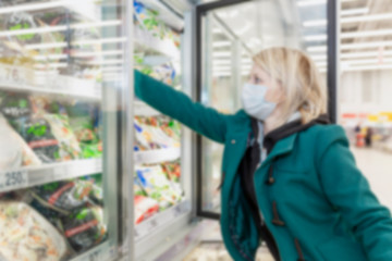A blond woman in a medical mask chooses products in the freezing department in a supermarket. Precautions during the coronavirus pandemic. Blurred.