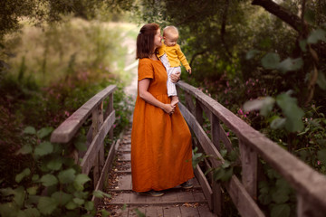 Fototapeta premium Caucasian mother with baby in her arms in a park on a wooden bridge, mother in a linen dress holds a one-year-old baby in her arms, walking in the park, concept motherhood and summer, walks near home