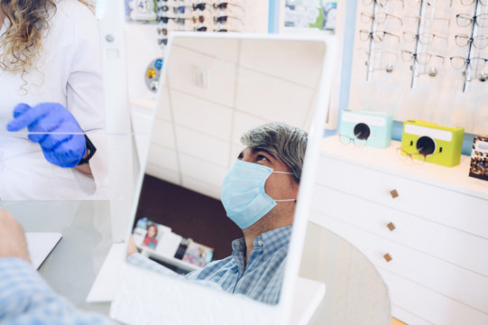 Gray-haired Man With A Face Mask Reflected In A Mirror While Talking To The Especialist Doctor In An Eyewear Store