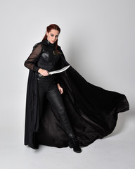 fantasy portrait of a woman with red hair wearing dark leather assassin costume with long black cloak. Full length standing pose holding a sword isolated against a studio background.