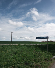 Electricity poles on green famlands next to a rural road and a street sign with the name of the nearby lake Svaneholmssjön on a bright spring day in southern Sweden