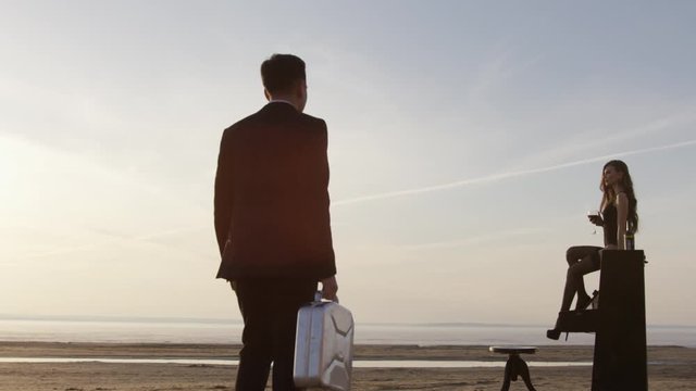 Beautiful young woman with long hair sits on a piano on the beach at sunset, a man in a black suit is heading towards her. Art composition