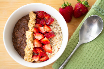 Chocolate cocoa oatmeal with almond meal, chopped strawberries and creamy peanut butter on a wooden surface. Strawberry, almonds, napkin and spoon in the background