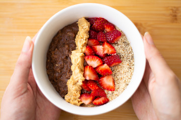 Chocolate cocoa oatmeal with almond meal, chopped strawberries and creamy peanut butter on a wooden surface. Strawberry, almonds, napkin and spoon in the background