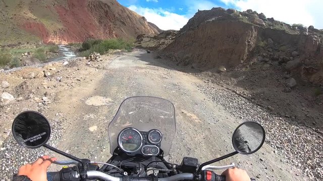 A wide point of view shot of slow riding a bike on a high altitude Himalayan mud road in bright daylight. Bike is driving through colourful scenic, barren mountain ranges of Jammu Kashmir region