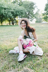 Fototapeta premium Portrait of a young woman in a hat and an armful of peonies. Beautiful stylish girl in a summer dress, a hat and a bouquet of flowers. Girl and flowers.