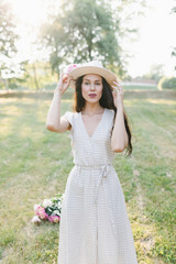 Portrait of a young woman in a hat. Beautiful stylish girl in a summer dress, a hat and a flower in her hand.