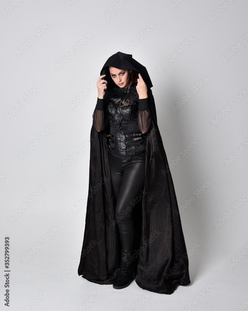 Poster fantasy portrait of a woman with red hair wearing dark leather assassin costume with long black cloak. Full length standing pose  isolated against a studio background.