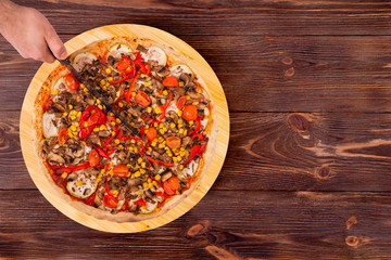 Man cut the pizza with a kitchen knife. Pizza with mushrooms, corn, cherry tomatos, courgettes and bell peppers on the wood platter which is on wooden rustic table, top view and copy space