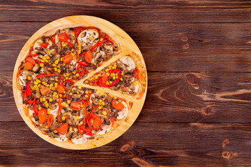 Pizza with mushrooms, corn, cherry tomatos, courgettes and bell peppers, with a slice removed on a wood platter which is on wooden rustic table, top view and copy space