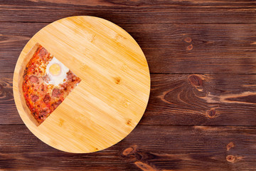 A quarter of tasty pizza with beef sausages, eggs and bacon on a wood platter which is on wooden rustic background, top view and copy space