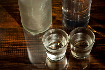 Frozen glasses with alcohol near the bottle. Drinks on a dark brown table. Transparent utensils on a black background. Old shabby boards under glass.