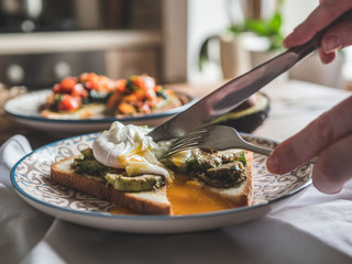 Close-up view cutting perfect poached eggs with avocado paste on crispy toast, fresh green avocado spread sandwiches recipe in restaurant as healthy breakfast.