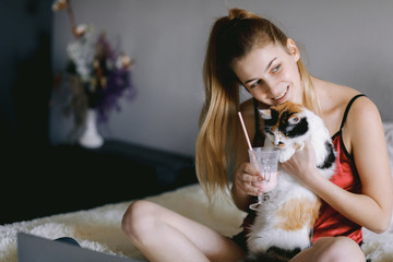 Young woman in silk sleepwear sipping milkshake and playing with cat during break in studies on bed at home