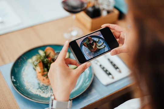 Young beautiful woman enjoying in tasty and nicely decorated meal. She sitting in expensive restaurant and using her smart phone for food photography.