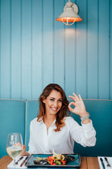 Young beautiful and happy woman enjoying in delicious meal in luxurious restaurant. She is smiling and showing ok sign.