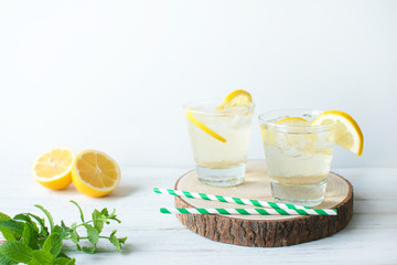 Two glasses of lemon homemade lemonade with mint leaves and ice on round wooden cut board on light background. Cool and fresh detox drink. Space for text.