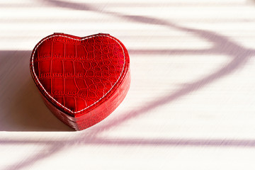 Red heart-shaped jewelry box made of leather on a white wooden background, natural sunlight, hard shadows. Gifts for lovers. Valentine's day. Copy space. The concept of love and luxury