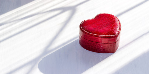 Red heart-shaped jewelry box made of leather on a white wooden background, natural sunlight, hard shadows. Gifts for lovers. Valentine's day. Copy space. The concept of love and luxury