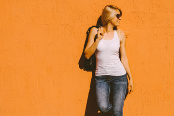 young  smiling blonde woman in white tank top and sunglasses against yellow wall outdoor in city...
