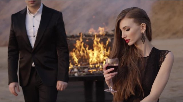 A young beautiful woman with a make-up and long hair on a sand quarry sits with a glass of red wine, a man in a classic black suit passes by. In the background is a flaming piano
