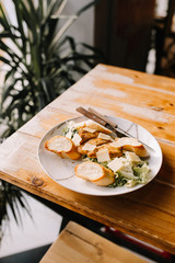 Caesar salad in a beautiful plate stands on a wooden table in a cafe on a sunny day. Caesar with chicken and white bread croutons with parmesan and anchovy dressing.