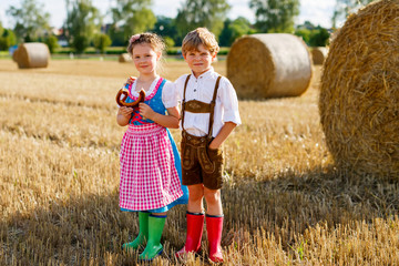 Two kids in traditional Bavarian costumes in wheat field. German children eating bread and pretzel during Oktoberfest. Boy and girl play at hay bales during summer harvest time in Germany.