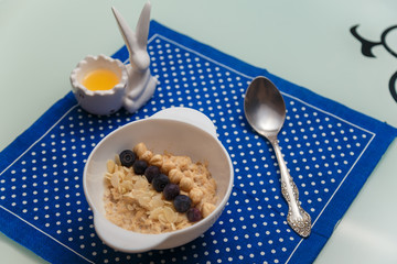 White bowl of oatmeal porridge with blueberries, almond flakes, nuts. Pot of honey and spoon on blue napkin on kitchen table. Healthy eating habits and diet concept.