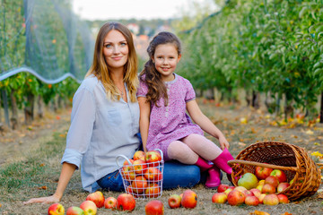 Portrait of little girl and beautiful mother with red apples in organic orchard. Happy woman and kid daughter picking ripe fruits from trees and having fun. Harvest season for family.