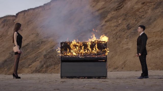 A man and a woman stand opposite and look at each other, between them a piano burns with a bright flame. scene in a sand pit. Shows the relationship. Art production, art scene.
