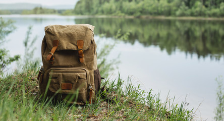 Obraz premium Tourist's brown backpack on the shore of a river. A great trip to nature in a dense green forest with a reservoir